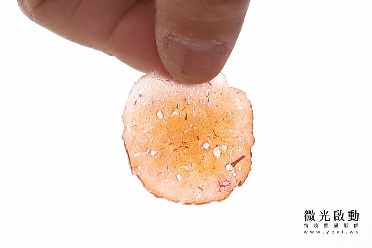 一隻手捏著一片半透明、帶有白色點狀顆粒的橘色圓形寵物零食，背景為純白色。