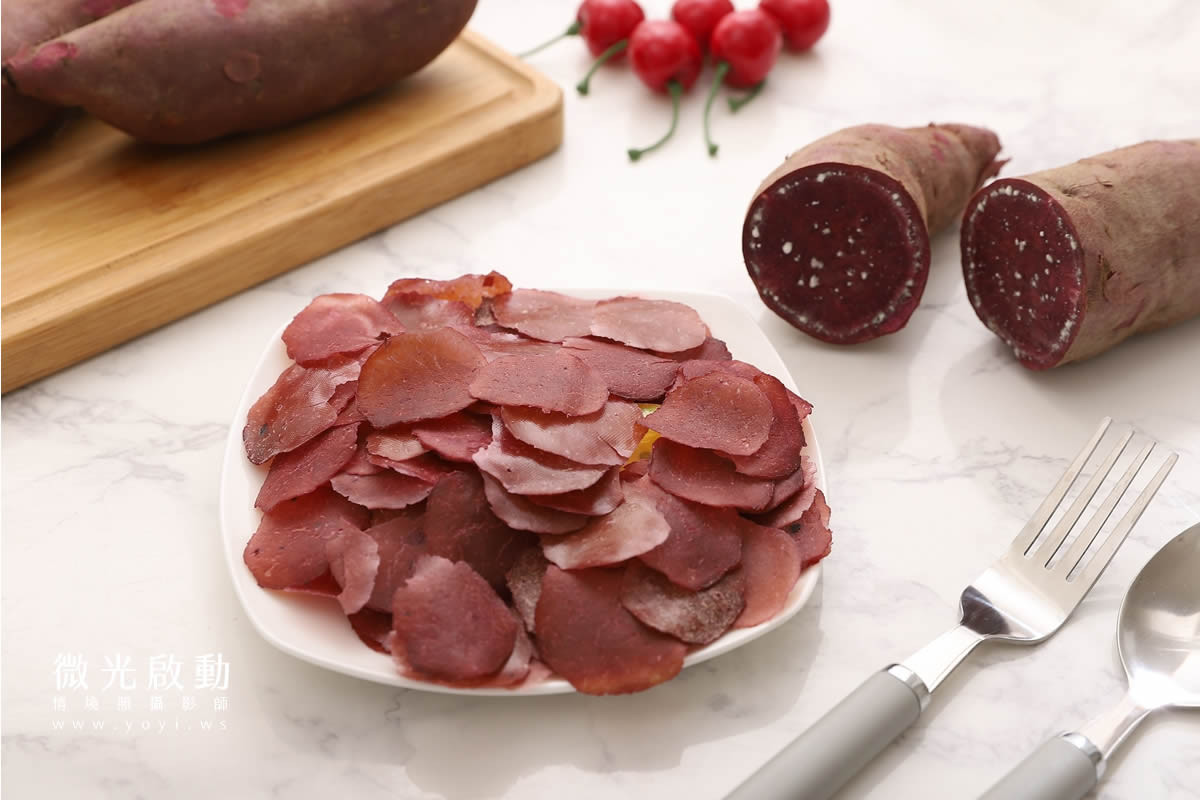 一盤深紅色圓形寵物零食，背景擺放著整根及切半的紫色地瓜，還有幾顆紅色小番茄，畫面呈現天然食材的健康感。