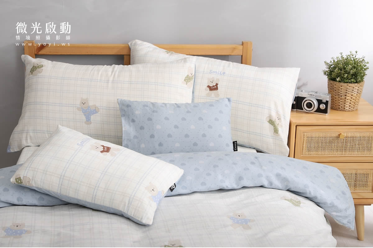 寢具攝影作品。淺藍色和白色格紋的棉被與枕頭組，在木質床架上整齊擺放，展示床組的細節與色彩。強調商品攝影、攝影服務。