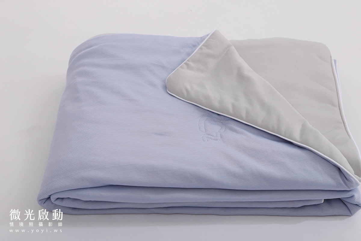 攝影專案作品。一張淺藍色條紋的棉被，其柔軟的折疊與精緻的刺繡細節在白色背景下清晰可見。標示寢具攝影、商品攝影、專業攝影。