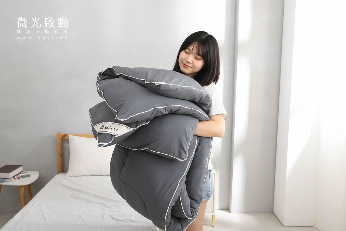 專業攝影服務作品。一位模特兒抱著折疊後的灰色棉被，展現其蓬鬆與輕盈感。適用於居家商品攝影、寢具攝影。