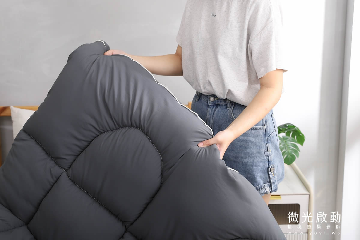 專業攝影作品。深灰色棉被在簡潔的臥室中，搭配白色床單，營造出對比與層次感。標示寢具攝影、居家商品攝影。