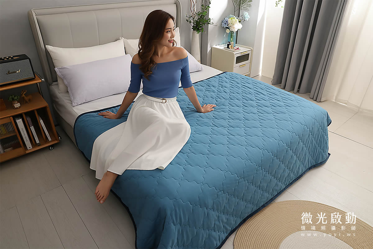 亞洲女性看起來大約20多歲，擁有棕色的長髮。她的妝容精緻，臉上帶著溫暖的微笑。她穿著一件藍色露肩上衣和一條白色長裙，腰間繫著一條白色細皮帶，眼神看著窗外她雙腳蜷曲著坐在床上，身體向窗戶的方向轉