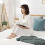 淺色寢具攝影，舒適居家風格，女模特兒穿著白色睡衣坐在床上閱讀，畫面中有木質床架、抱枕與毛毯。