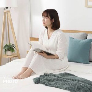 淺色寢具攝影，舒適居家風格，女模特兒穿著白色睡衣坐在床上閱讀，畫面中有木質床架、抱枕與毛毯。