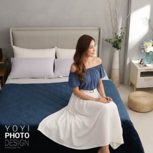 專業寢具攝影作品：深藍色菱格紋床包，搭配灰色與白色枕頭，模特兒身穿藍色上衣與白色長裙，坐在床邊，展示高質感居家寢具生活風格。