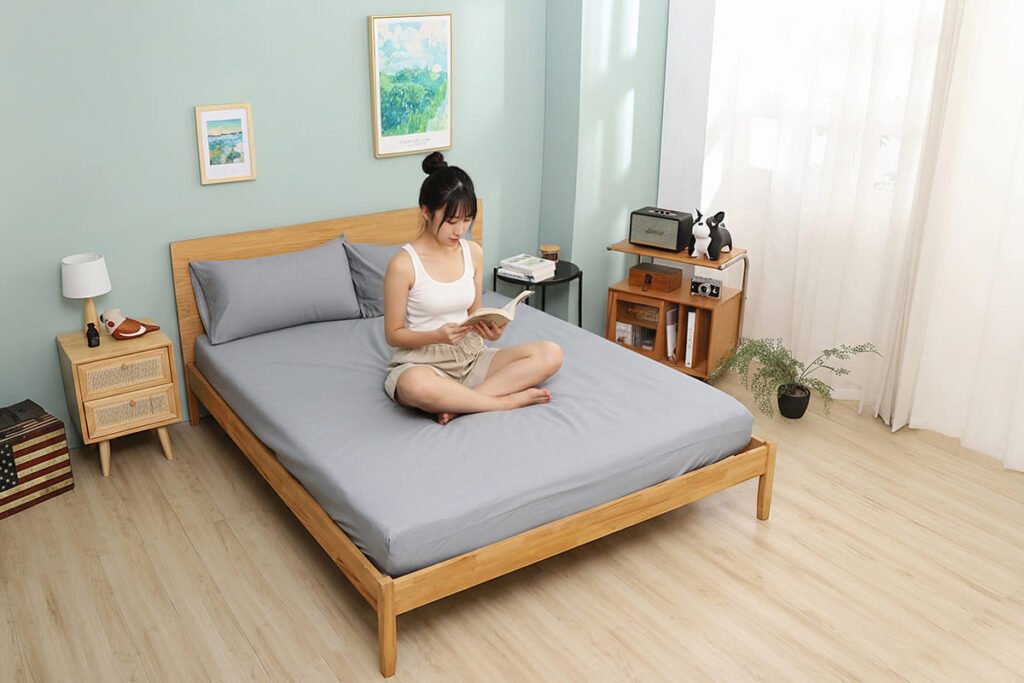 淺灰色純棉床包組搭配原木簡約雙人床架的情境攝影。一位女性模特兒穿著休閒服，坐在床上閱讀，展示了床單的柔順質感和舒適居家生活情境。適用於現代簡約風臥室設計。